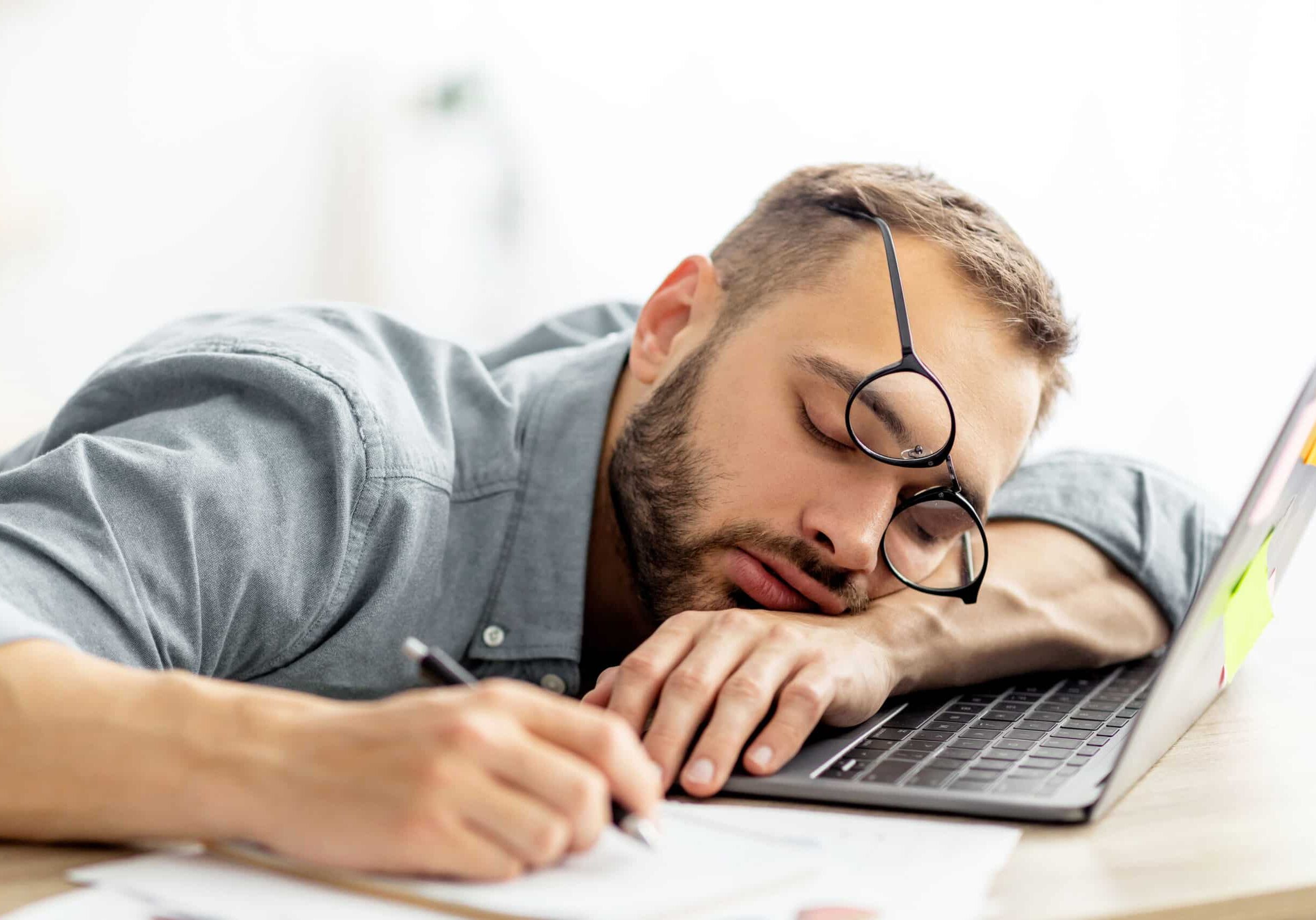 Exhausted millennial man sleeping on his office desk, next to laptop and documents, tired of overworking. Young male workaholic suffering from chronic fatigue at workplace