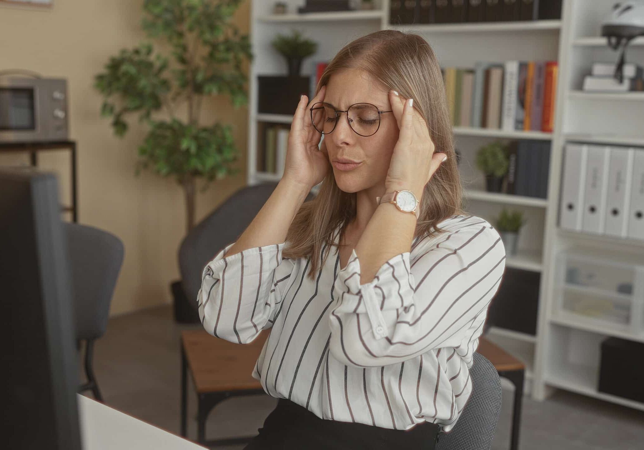 Woman wearing glasses and striped blouse pressing temples next to computer monitor inside office building; stress.