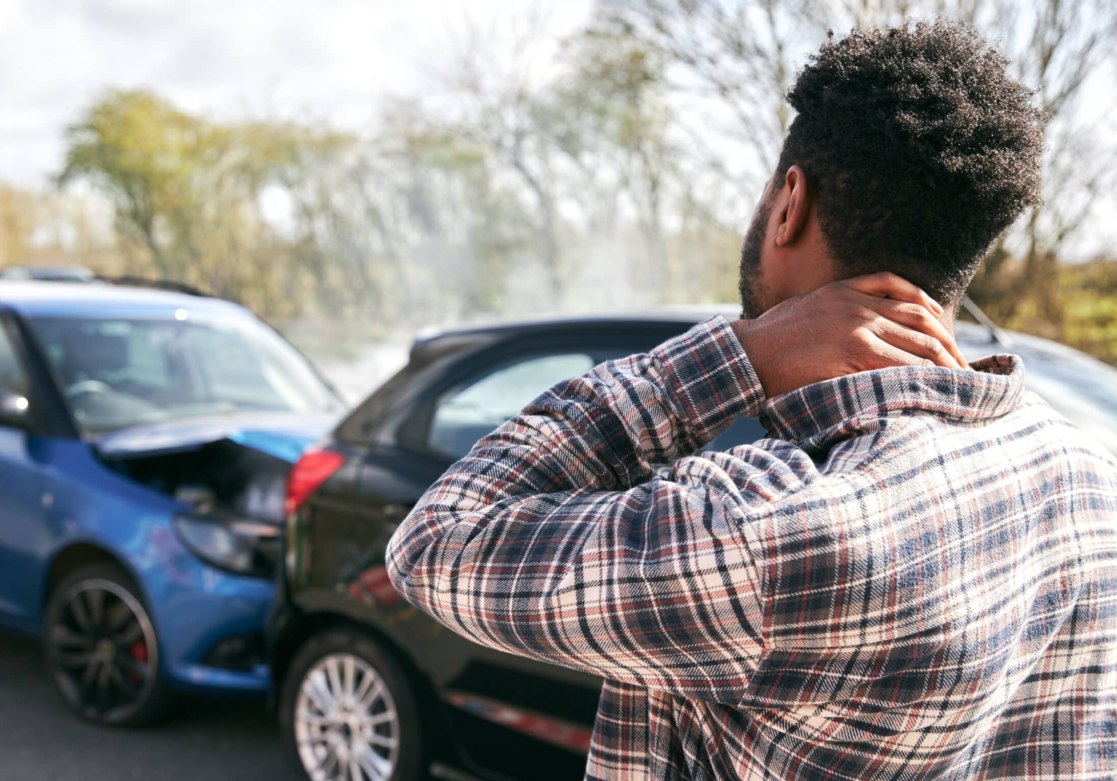 Young man rubbing neck in pain from whiplash injury standing by damaged car after traffic accident